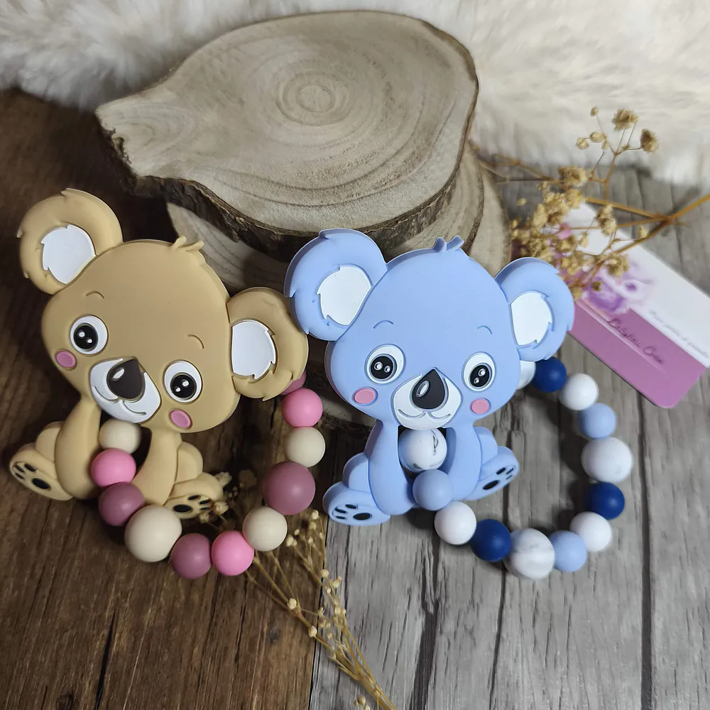 Deux anneaux de dentition en silicone modèle koala, l'un beige avec des perles roses et crème, l'autre bleu avec des perles bleues et blanches, présentés côte à côte sur un fond en bois avec une décoration naturelle. Babybou-Créa