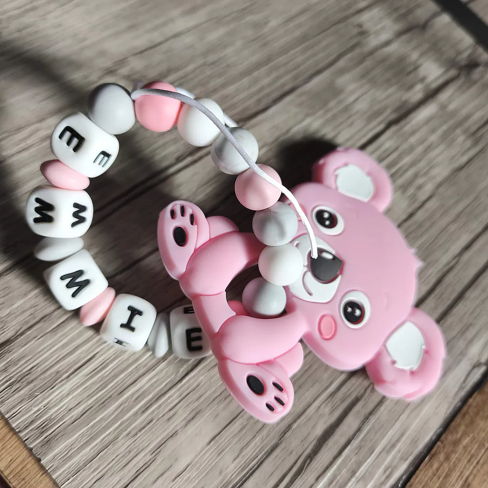 Anneau de dentition personnalisé en silicone avec un koala rose et des perles roses, blanches et effet marbre. Le prénom « EMMIE » est intégré aux perles lettres. La création est photographiée sur un fond en bois. Babybou-Créa