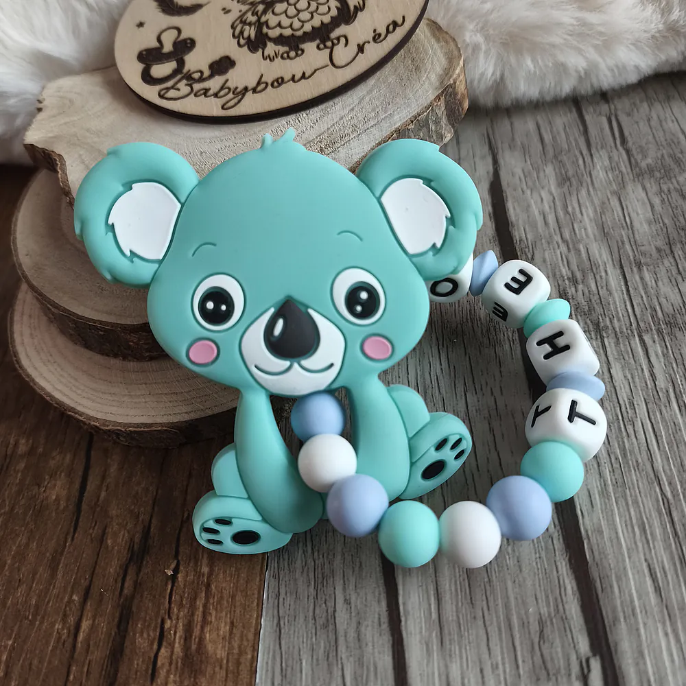 Anneau de dentition personnalisé en silicone avec un koala turquoise et des perles bleu pastel, turquoise et blanches. Le prénom « THEO » est intégré aux perles lettres. La création est photographiée sur un fond en bois naturel. Babybou-Créa
