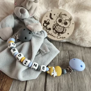 Attache doudou personnalisée en silicone avec deux clips gris et bleu, motif renard, perles grises, bleues et moutarde, personnalisée avec le prénom Ondine, présentée avec un doudou.